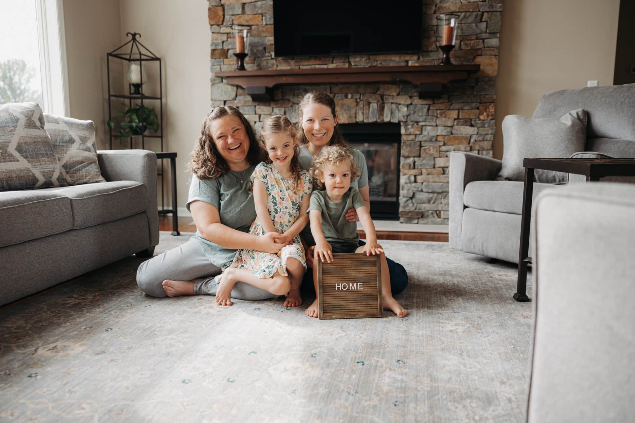 Family of four smiling together in a cozy living room, holding a sign that says "HOME," representing the warmth and connection of homeownership, relevant to First Class Mortgage's mission for Minnesota homebuyers.